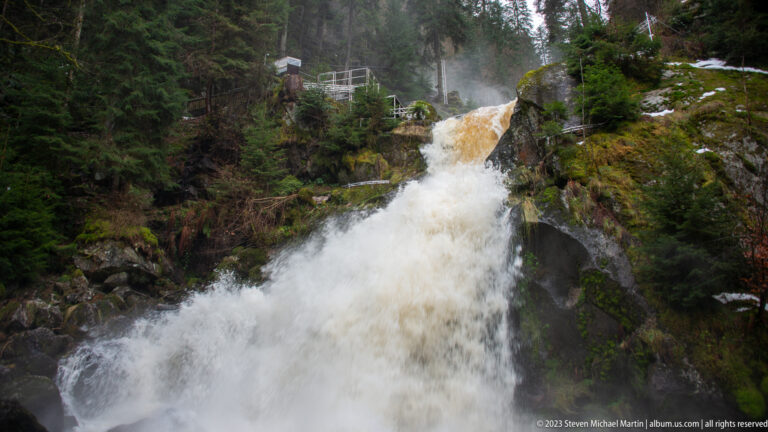 Powerful Triberg waterfall plunging between dark mossy rocks, with spray, snow patches, and a railing above.