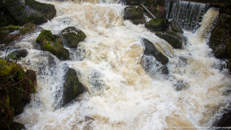 Foaming Triberg waterfall water rushing around dark mossy boulders, with a smaller curtain of water at upper right.
