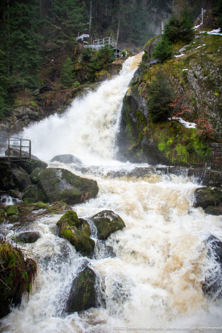 Broad Triberg cascade flowing over rock shelves into churning water, framed by forest, snow, boulders, and railings.