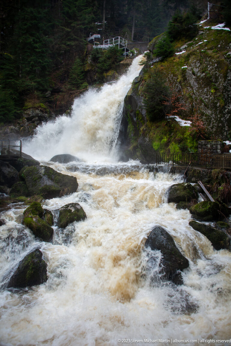 Low broad cascade at Triberg Waterfalls rushing across dark boulders, with forest and a visitor railing behind.