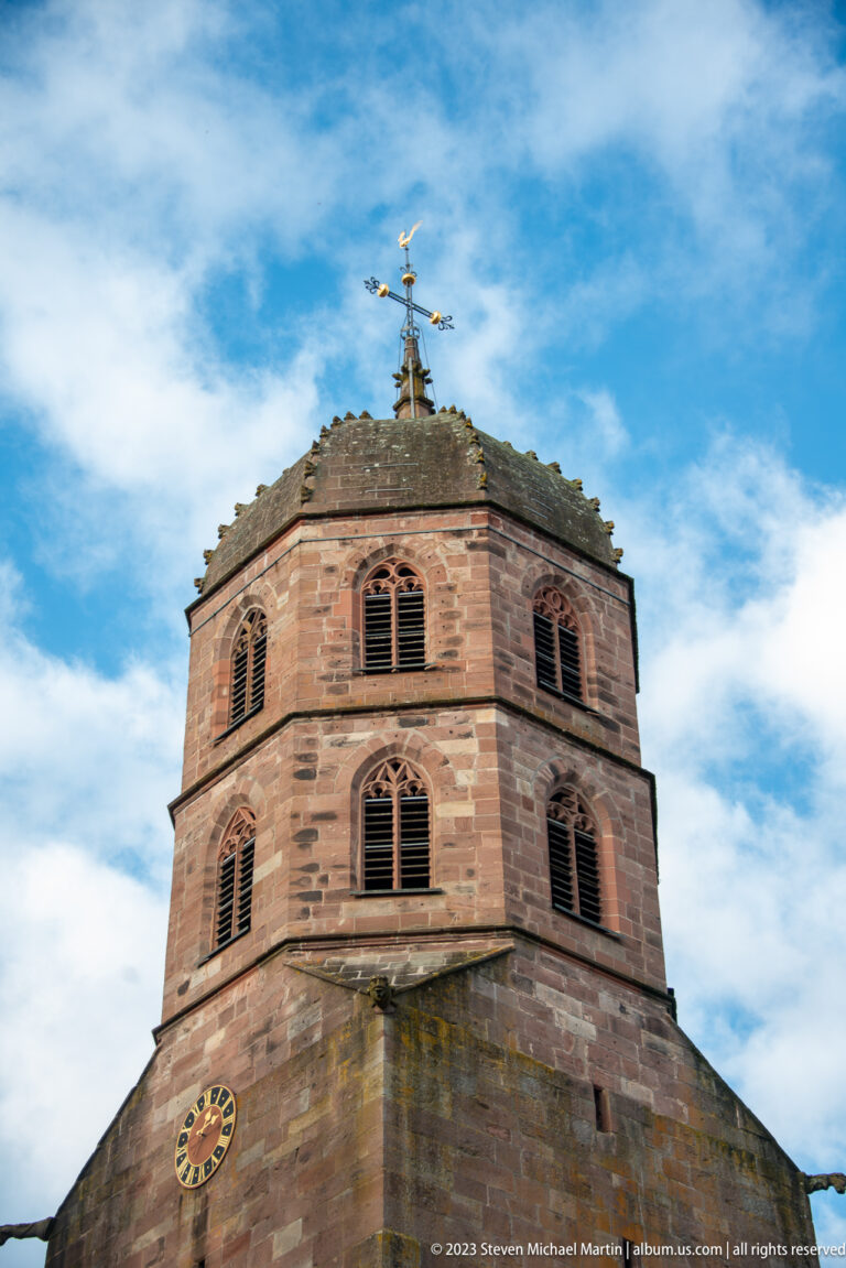 Octagonal reddish sandstone church tower with pointed-arch louvers, small clock, low roof, and metal finial.