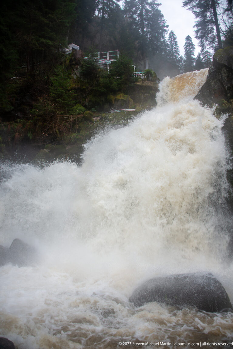 Wide Triberg cascade surging through misty spray, with dark rocks, forest, and a visitor railing above.