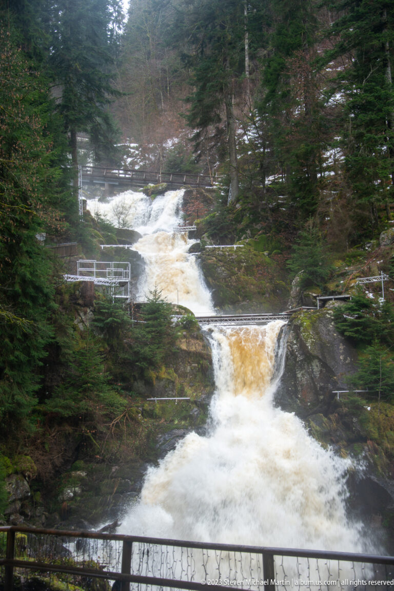 Multi-tier forest waterfall rushing beside railings and a footbridge among dark evergreen trees.