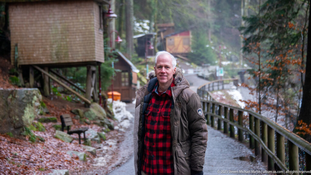 Visitor standing on a wet forest walkway beside a wooden railing, with snow and rushing water behind.
