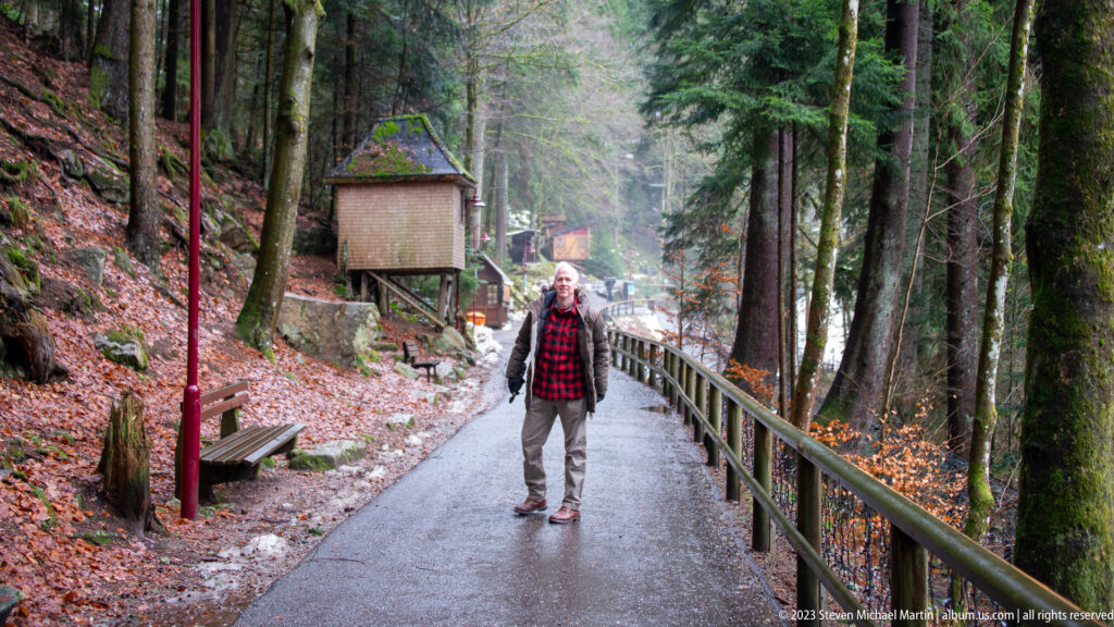 Visitor walking along a paved forest trail with benches, railings, small structures, and patches of snow.