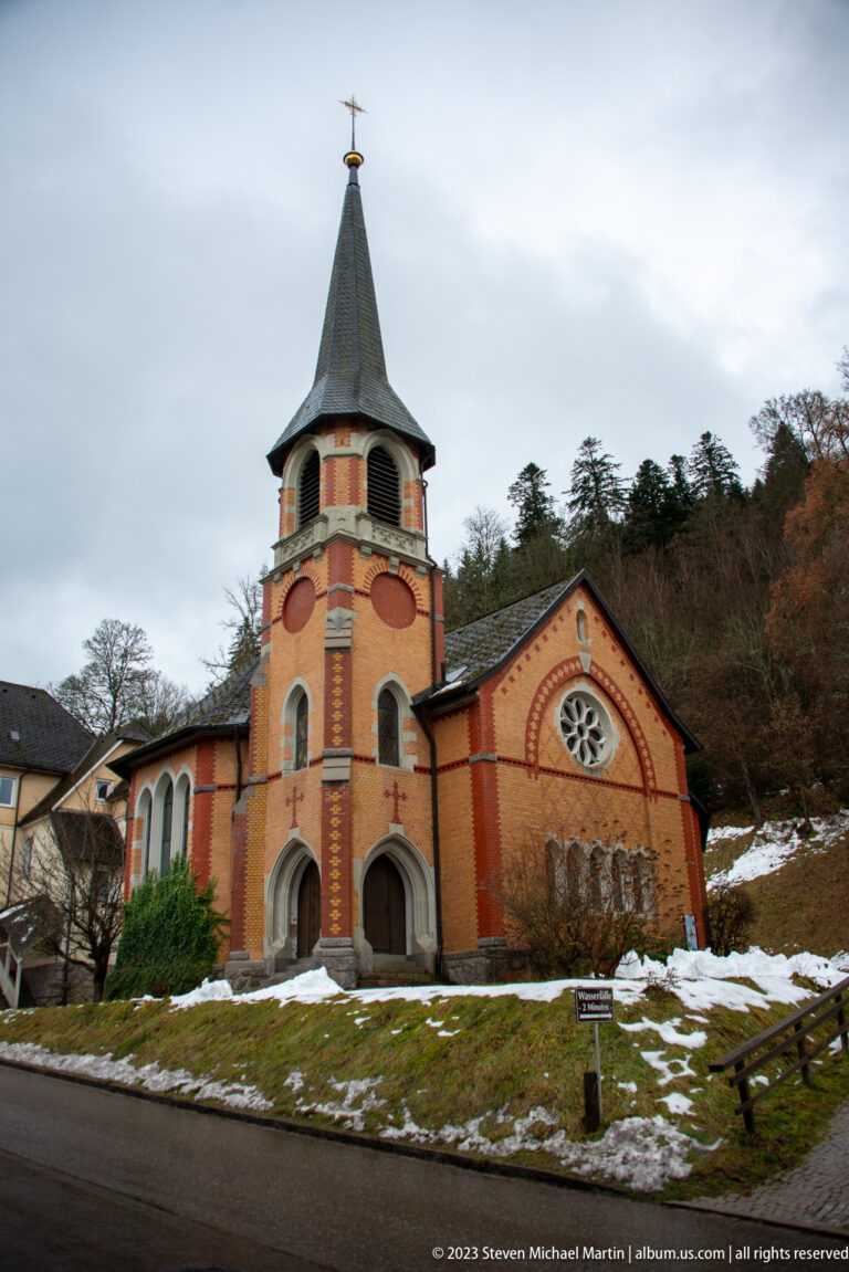 Brick church with square bell tower, steep roof, rose window, and snow patches on a wooded hillside.