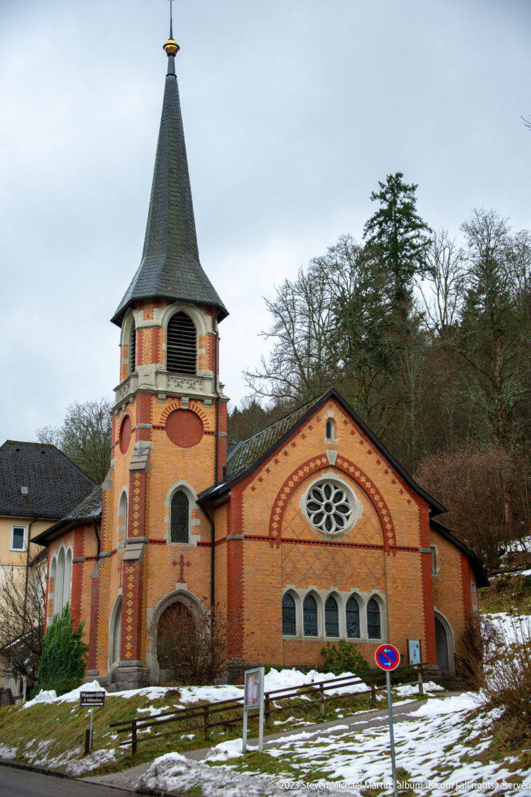 Front view of a brick church with steep bell tower, rose window, arched openings, and snow.