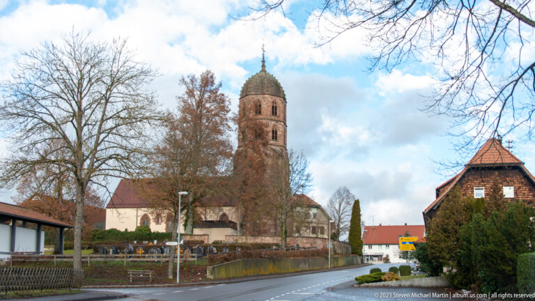 Village church with tall reddish bell tower, bare trees, houses, and a wet roadside in the Black Forest.