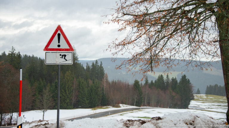 Warning sign beside a snow-lined road with conifer forest and hazy mountains beyond.