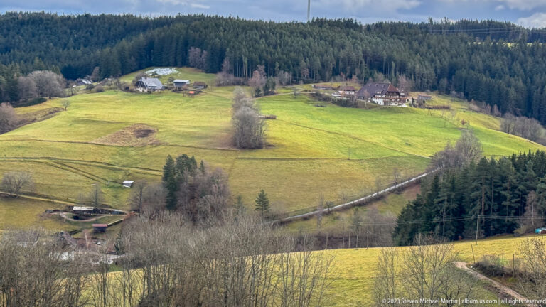 Elevated view over green rolling pastures, scattered farmsteads, leafless trees, and dark conifer forest.