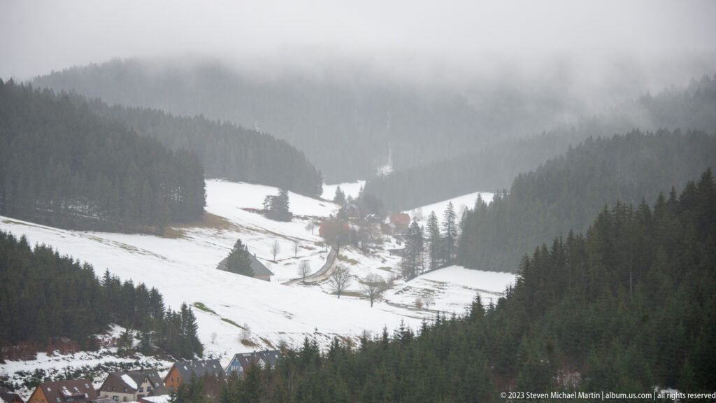 Misty snow-covered valley with dark conifer slopes, open meadows, road, and small settlement below.