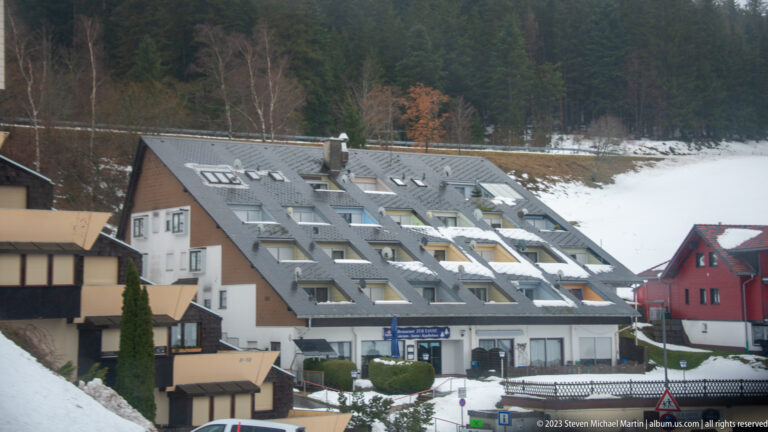 Large steep-roofed building with rows of dormer windows in a snow-covered Black Forest settlement.