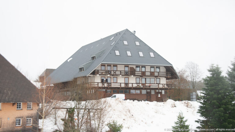 Large traditional Black Forest farm building with broad steep roof, timbered façade, balconies, and deep snow.