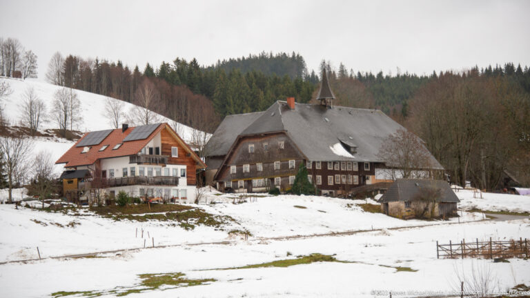 Traditional dark farmhouse with broad roof beside a newer chalet-style house on a snowy rural slope.