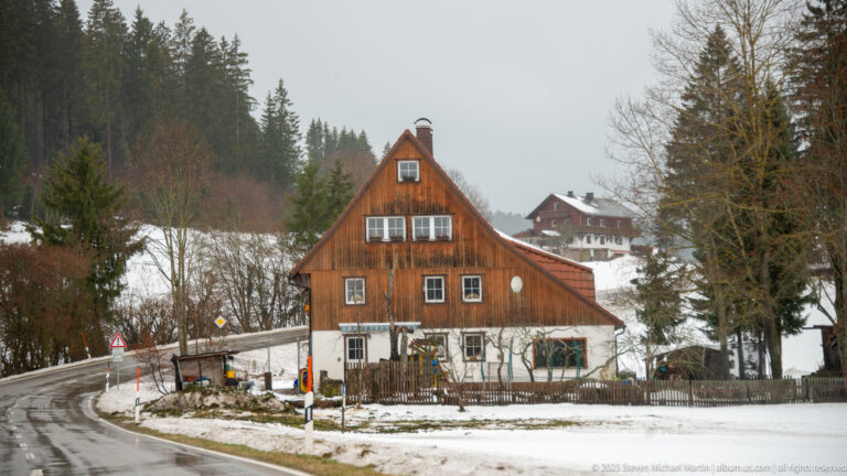 Timber-clad steep-roofed house beside a wet snowy road, with hillside buildings and forest behind.