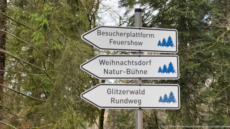 Three German directional signs in a forest point to a visitor platform, Christmas village stage, and Glitzerwald loop.