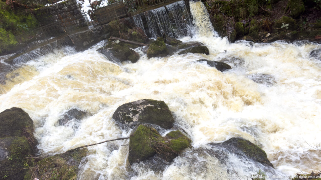Foaming water rushes over dark boulders below a low weir in a forested rocky channel.