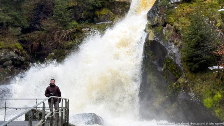 Large waterfall plunging beside a metal visitor platform where a single visitor stands for scale.