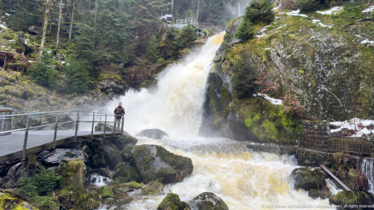 Broad forest waterfall behind a metal visitor walkway, with a visitor standing above the rocky outflow.