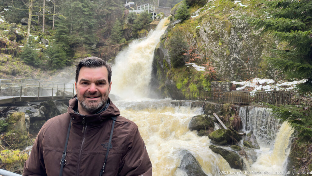 Visitor in a brown jacket standing before a foaming forest waterfall with mossy rocks, railings, and patches of snow.