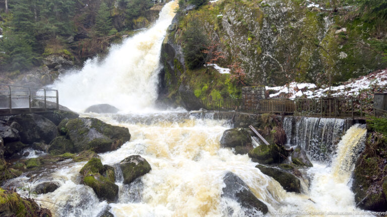 Powerful waterfall dividing around mossy boulders, with metal viewing railings and a smaller side drop.
