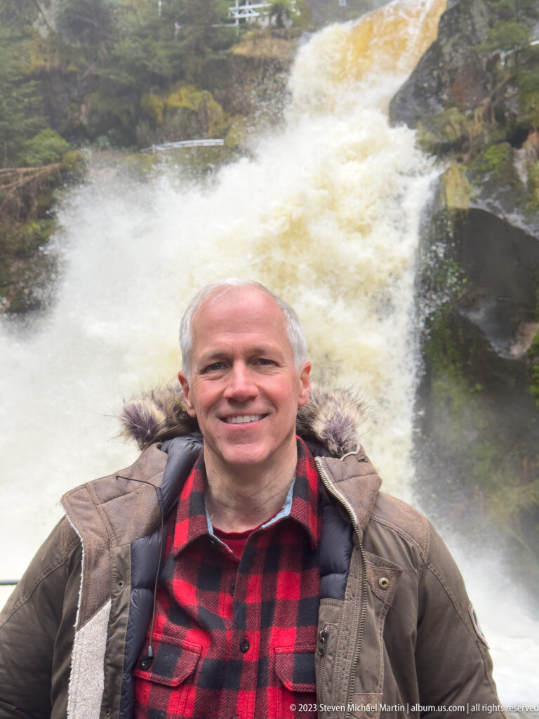 Smiling visitor in red-and-black plaid standing directly in front of a broad rushing waterfall.