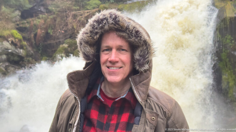 Hooded visitor in winter clothing standing close to a plunging waterfall beside dark mossy rock.
