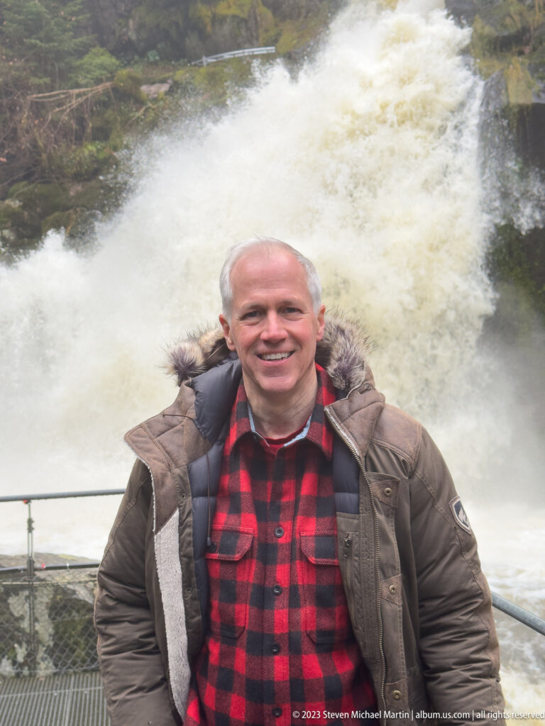 Visitor in plaid shirt and brown jacket standing before a broad foaming waterfall beside a metal railing.