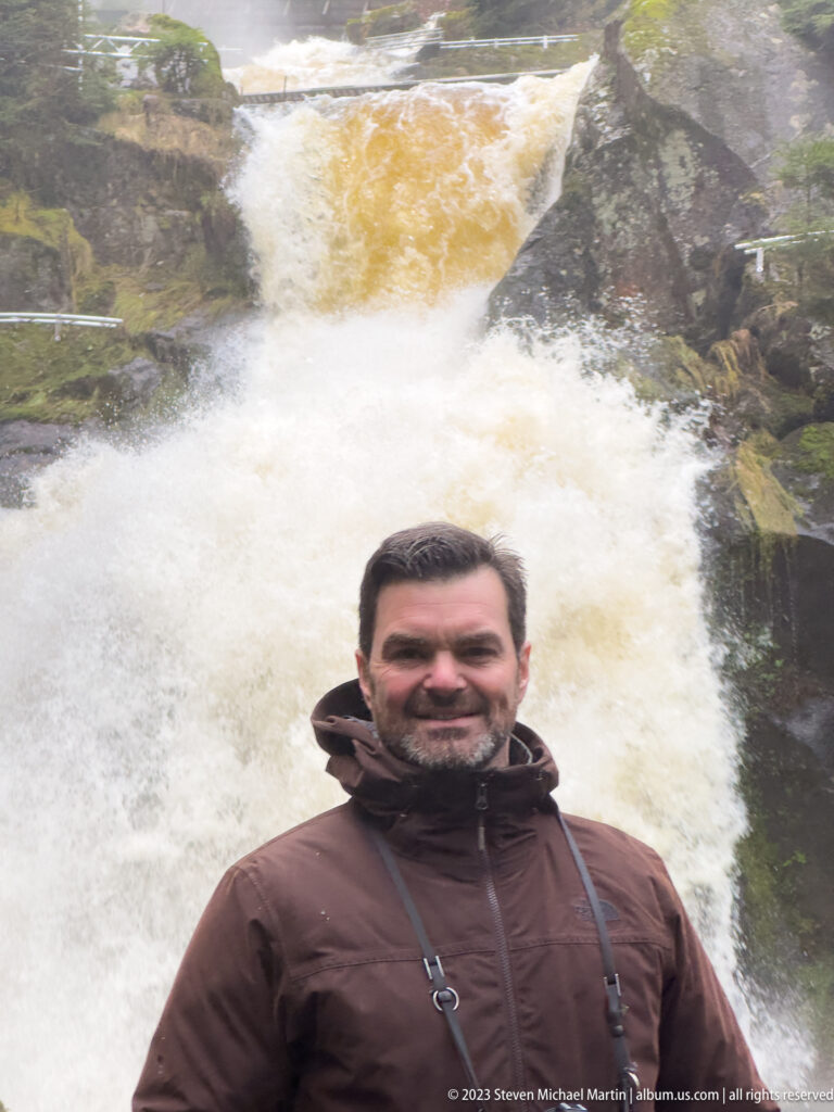 Bearded visitor in a brown jacket centered before a steep, turbulent waterfall filling most of the background.