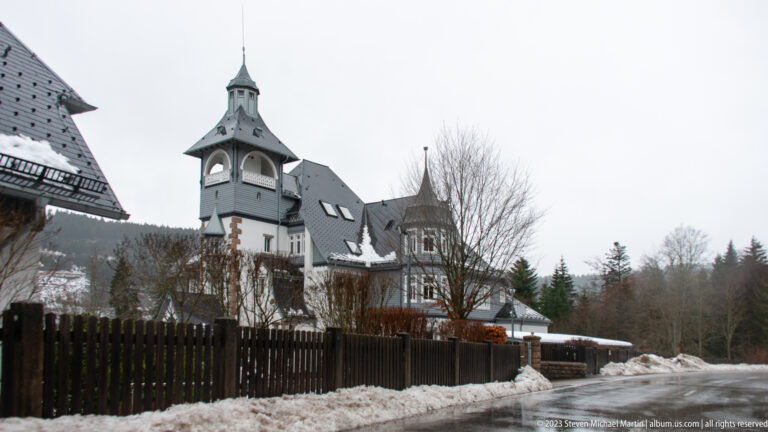 Ornate pale villa with square tower and steep roofs beside a road bordered by snowbanks and a dark fence.