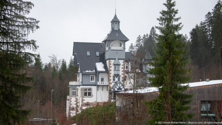 Towered white villa with steep dark roofs and masonry accents seen among evergreen trees and patches of snow.