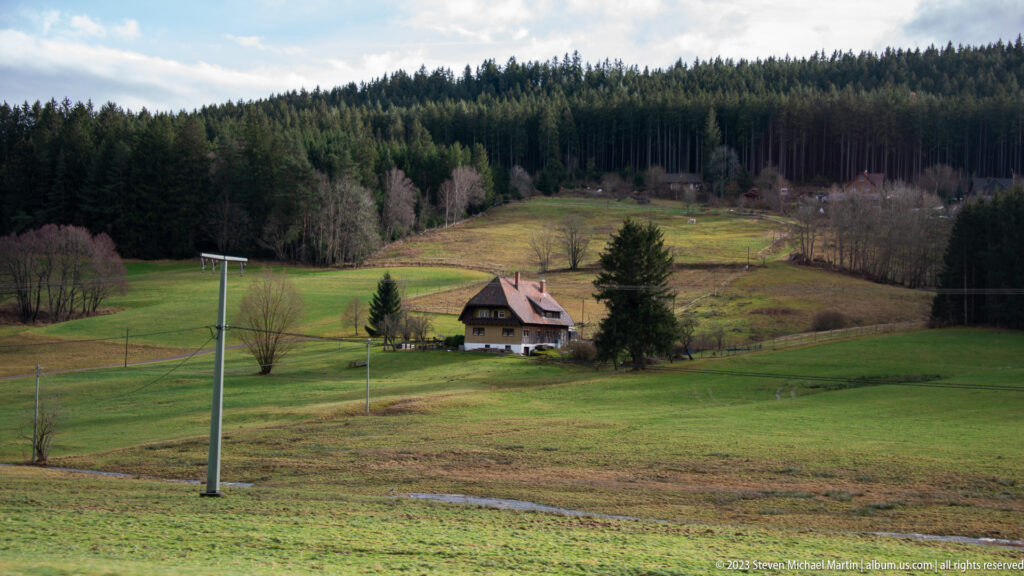 Farmhouse in green hillside meadows below dense conifer forest, with utility poles crossing the foreground.