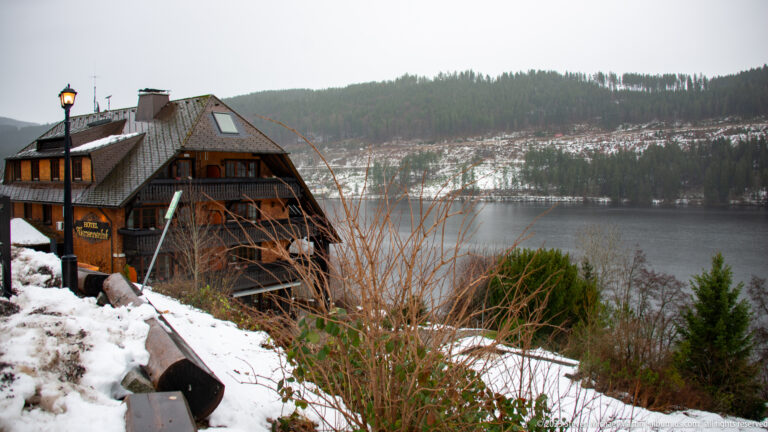 Large timber chalet with balconies on a snowy slope overlooking a lake and forested hills.