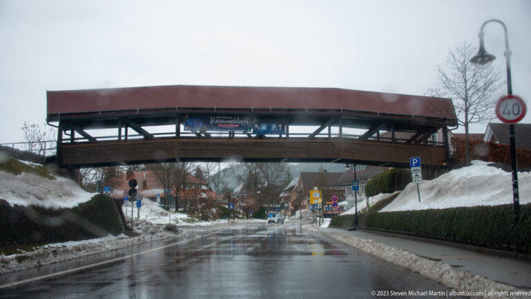 Covered timber pedestrian bridge crossing a wet road with snowbanks, traffic signs, and village buildings beyond.
