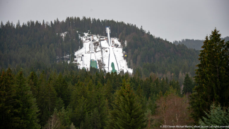 Snow-covered ski-jump ramps and tall structures descending through a cleared corridor in dense evergreen forest.
