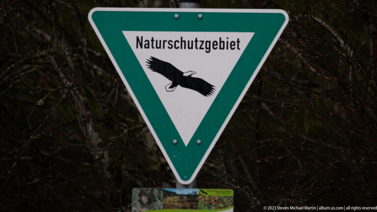 Green triangular Naturschutzgebiet nature-reserve sign with black bird silhouette against dense bare branches.