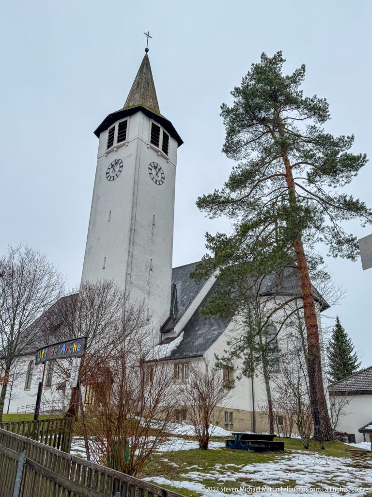White church with tall square clock tower and dark roof beside a large pine on a patchy snowy slope.