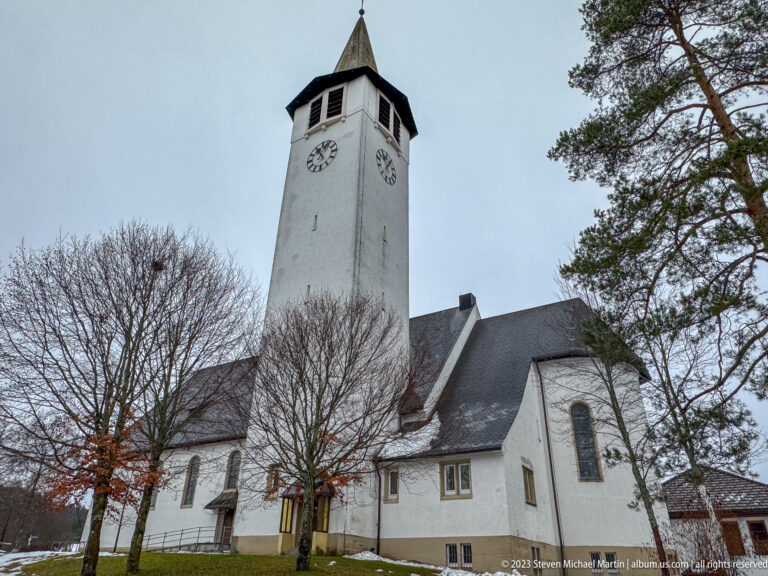 Low-angle view of a white church with square clock tower, dark roof, leafless trees, and patchy snow.