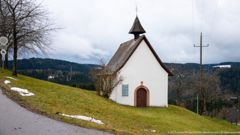 Small white hillside chapel with arched wooden door, steep roof, bell turret, and patches of snow.