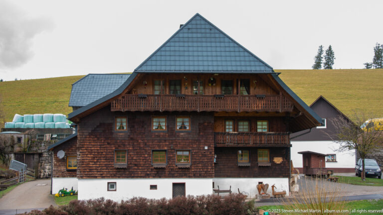 Large dark-shingled Black Forest house with two wooden balconies, steep roof, white base, and farm buildings behind.