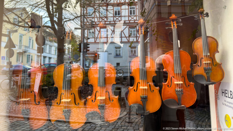 Violins displayed behind glass with reflections of half-timbered buildings and a cobbled street in Esslingen.