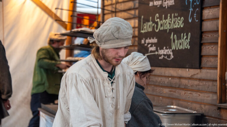 Young man in cream tunic and cap preparing food beside a handwritten chalkboard menu at a timber stall.