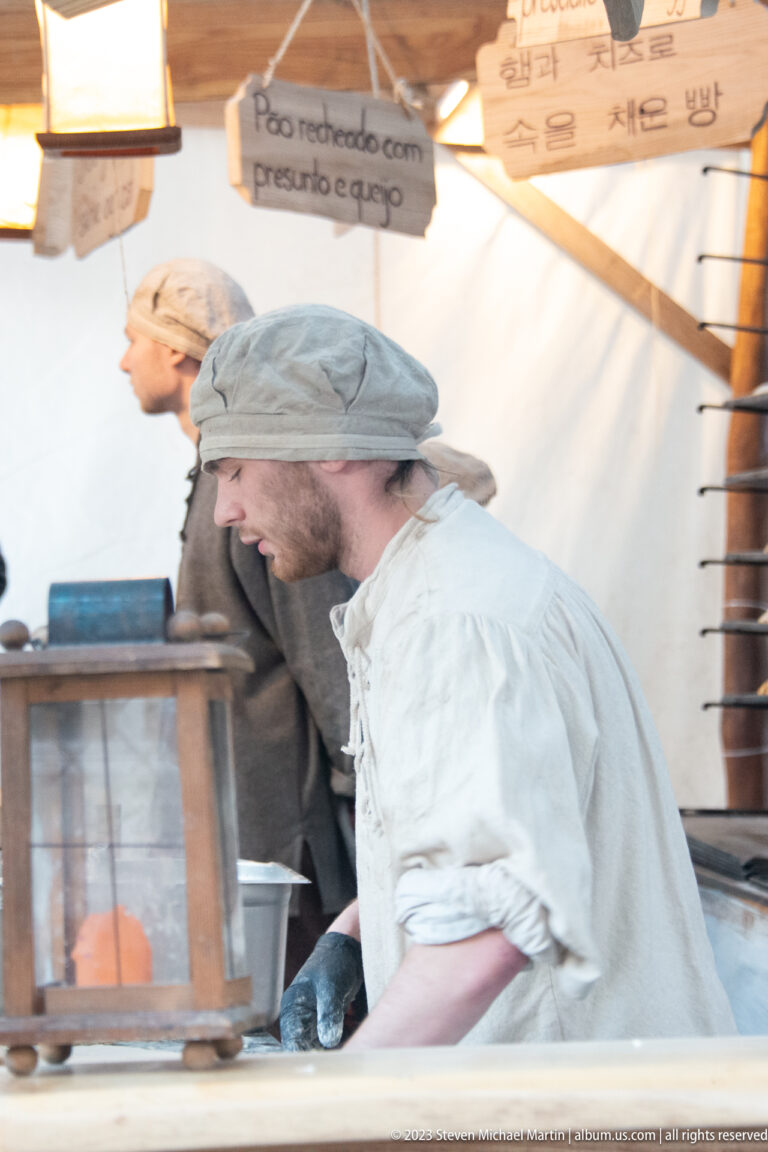 Costumed vendor in pale tunic leaning beside a box-like heated cooking vessel inside a market stall.