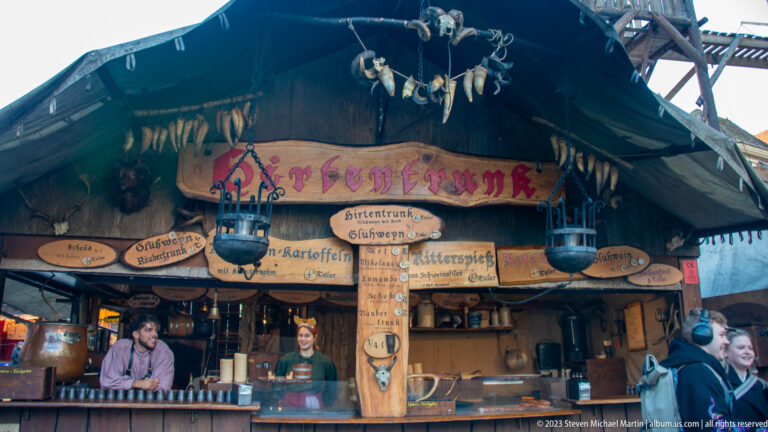 Rustic Kartoffelhaus food booth with painted menu boards, hanging garlic, pots, timber posts, and vendors.
