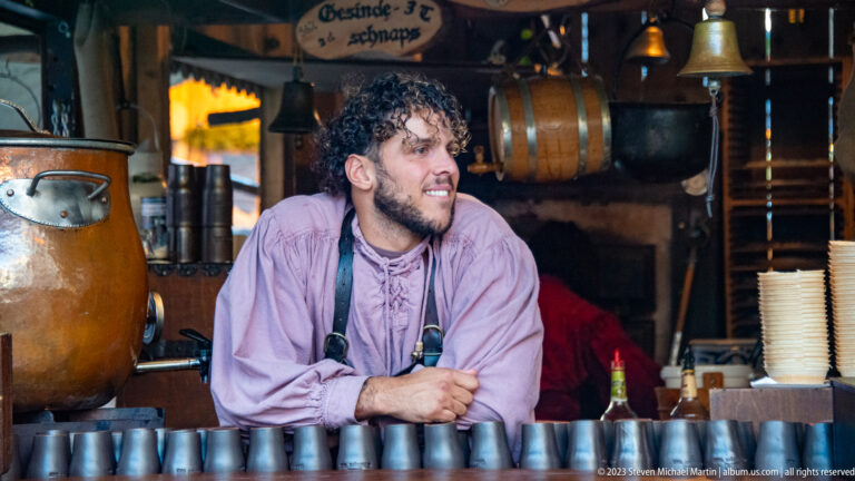 Smiling vendor in lavender shirt leaning behind metal cups, a copper vessel, hanging bells, and wooden barrels.