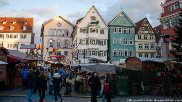 Christmas market crowd and timber stalls beneath colorful historic gabled houses, with part of the Altes Rathaus at right.