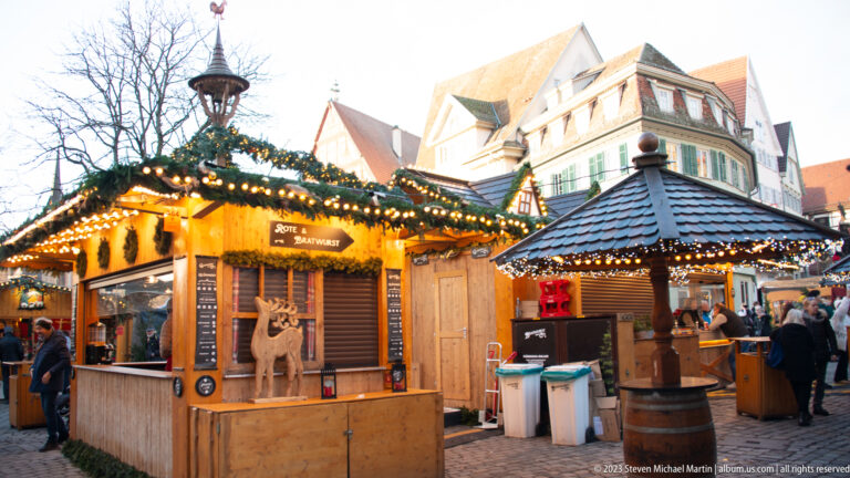 Two illuminated wooden Christmas market booths decorated with evergreen garlands and lights beneath historic buildings.