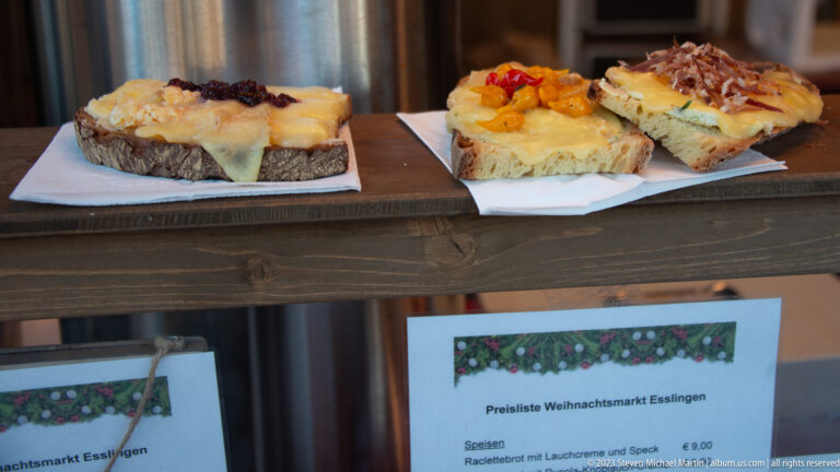 Open-faced rustic bread slices topped with melted cheese, peppers, and savory garnishes on a Christmas market counter.