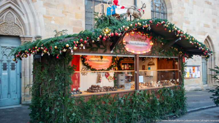 Evergreen-covered Christmas sweets stall with red ornaments and warm lights set against a pale stone church wall.