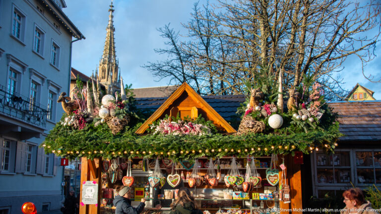 Wooden Christmas market stall with hanging heart ornaments, evergreen branches, flowers, baubles, and a Gothic spire behind.